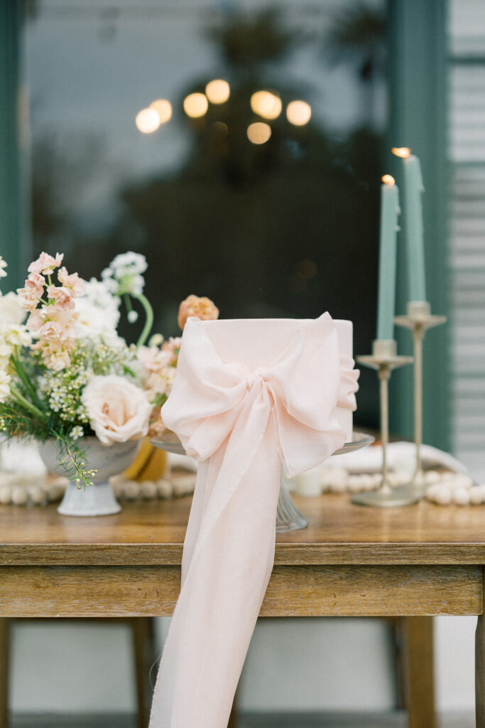 Pink wedding cake with pink bow around it on wood table with taper candles and flowers in the background.