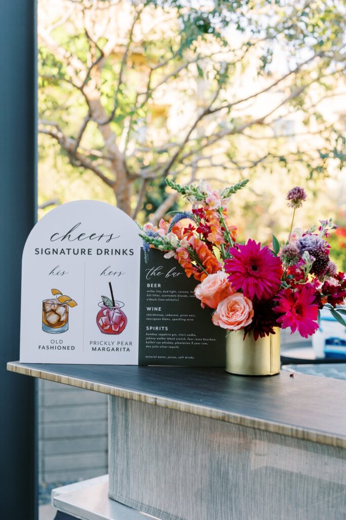 Wedding reception bar top with drink menu signs and small floral arrangement.