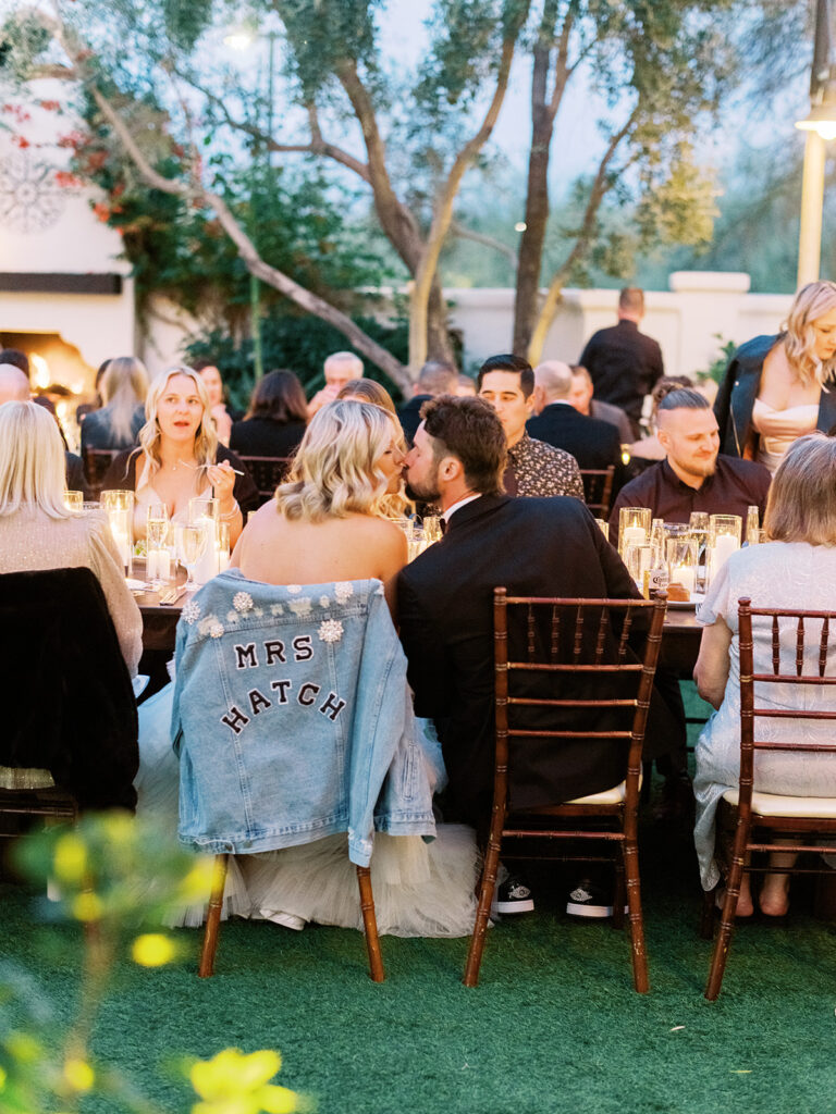 Bride and groom seated at reception table kissing, with denim jacket hanging over back of bride's chair.