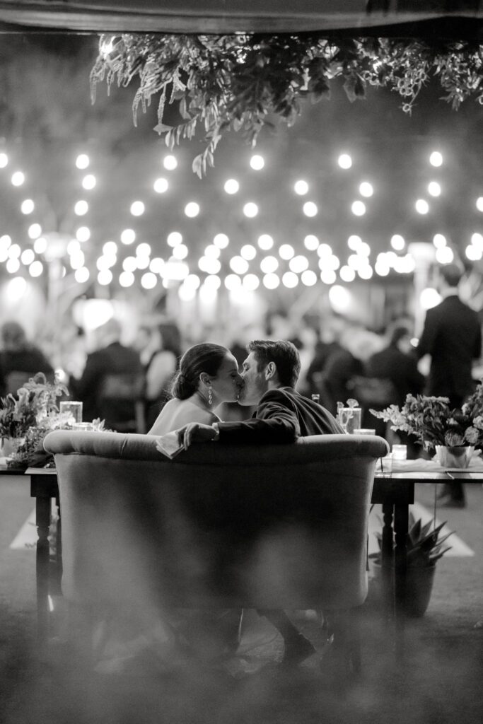 Bride and groom kissing on small couch behind sweetheart table.