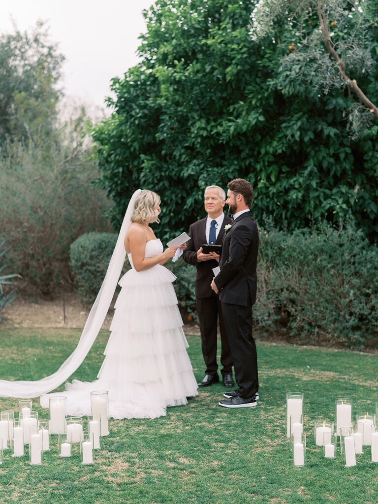 Bride reading vows to groom during wedding ceremony outside, with candles framing them in ceremony altar space.