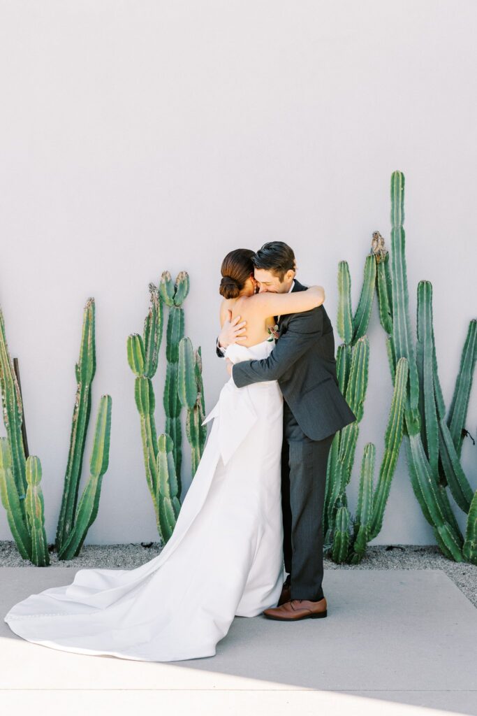 Bride and groom embracing in front of white wall with cacti plants growing in front of it.