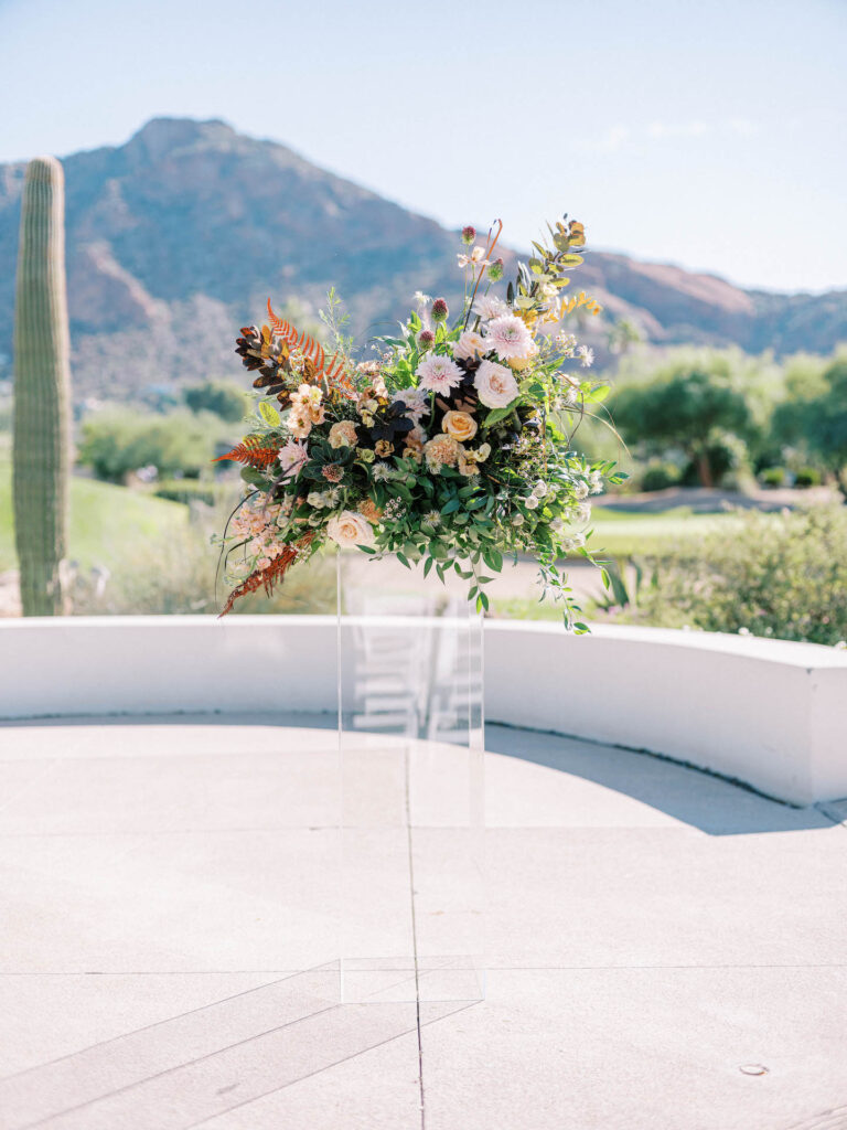 Wedding ceremony floral arrangment on clear acrylic stand of pink, peach, white flowers and greenery in orange and green.