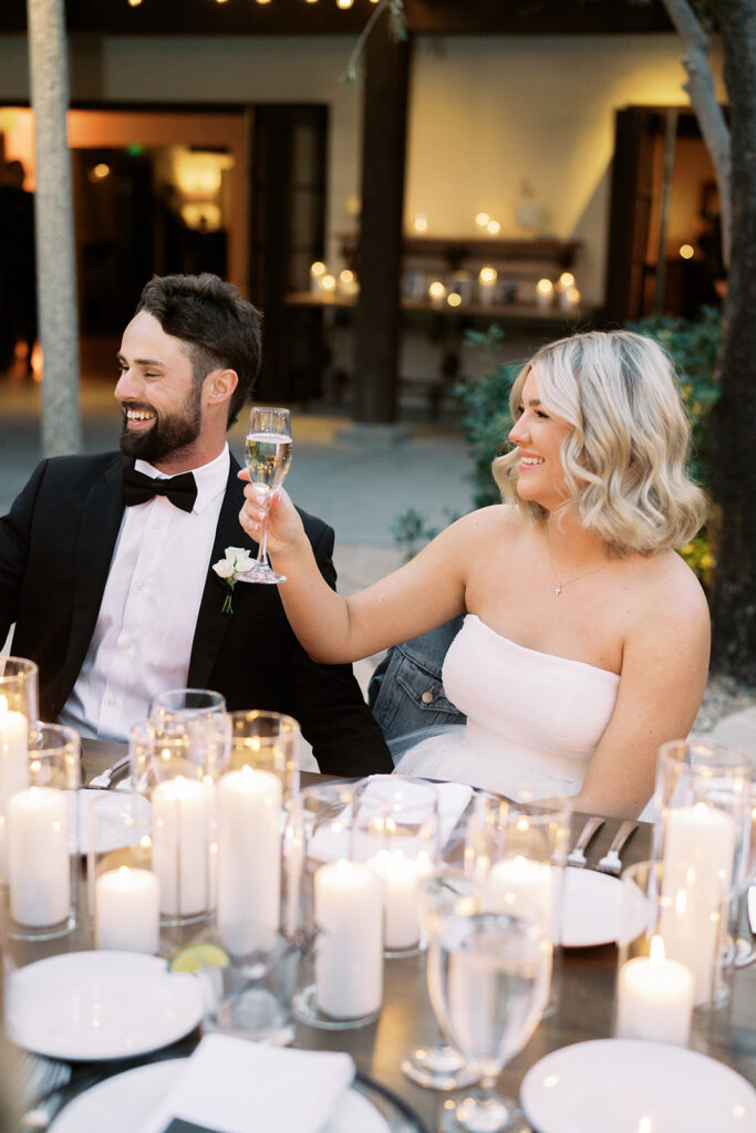 Bride and groom seated at wedding reception table smiling, bride holding up champagne glass in a toast.