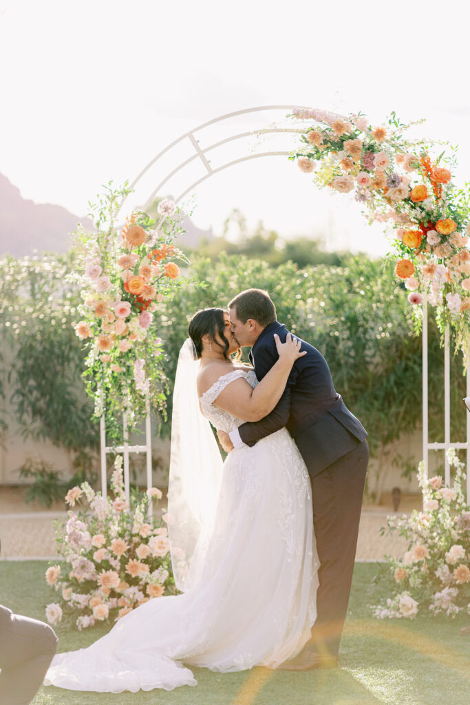 Bride and groom kissing under wedding ceremony arch with flowers installed on it.