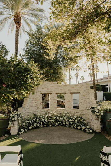 Outdoor wedding ceremony altar space with partial built white wall with large cutouts and floral of white on the ground along wall.