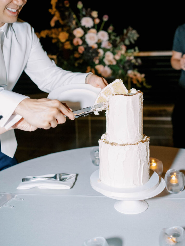 Groom removing a piece of cake from the top tier of a two tiered white wedding cake.
