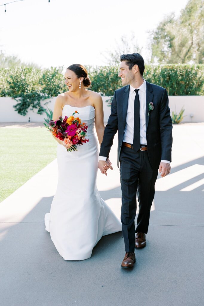 Bride and groom walking, holding hands, smiling, looking off to side.