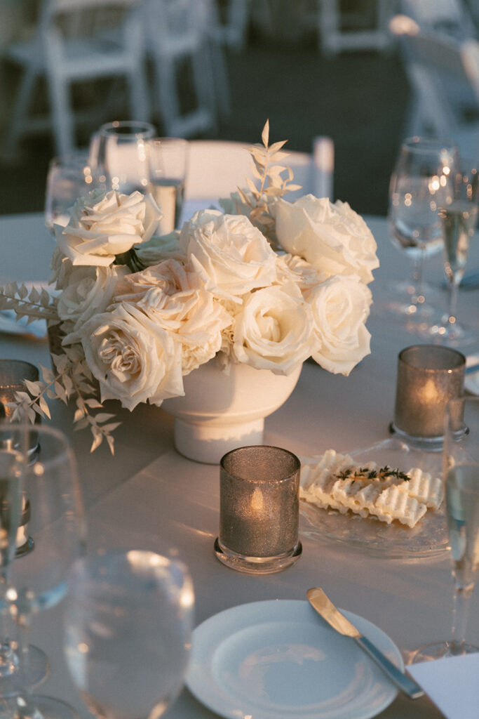 White roses and bleached Italian ruscus wedding reception centerpiece on round table with place settings and votive candles.