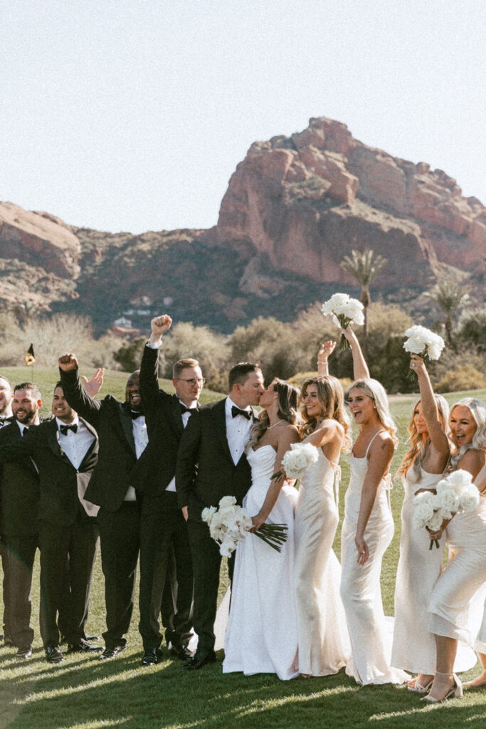 Bride and groom kissing between wedding party of groomsmen and bridesmaids celebrating with arms up in the air.