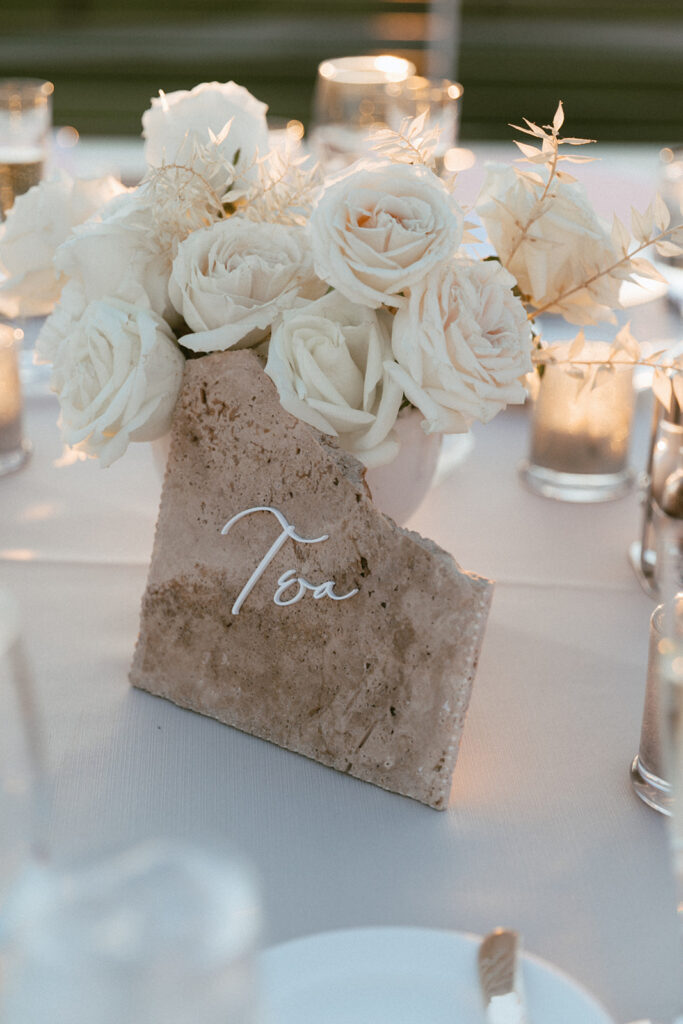 White roses and bleached Italian ruscus wedding reception centerpiece with stone table number sign in front.