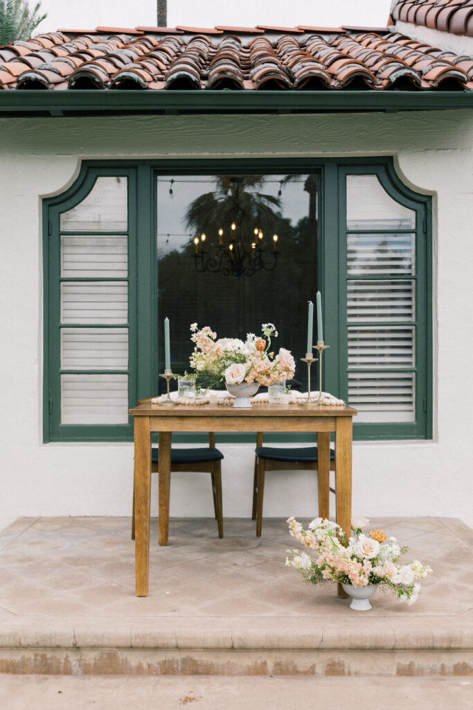 Wooden sweetheart table in front of house window with floral centerpiece on the top and on the ground with sage colored taper candles.