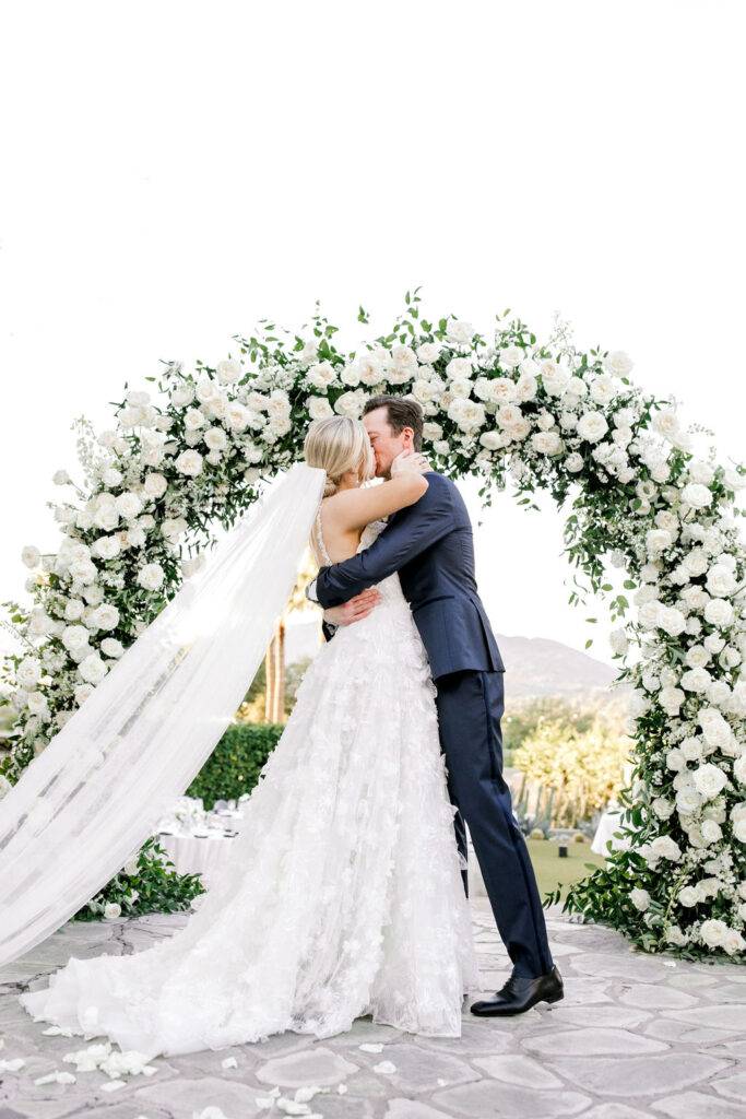 Bride and groom kissing and embracing in front of wedding ceremony arch of white flowers and greenery.