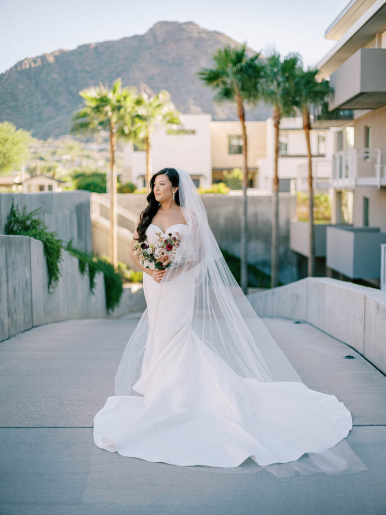 Bride standing on paved walkway holding a bouquet, looking off to side.