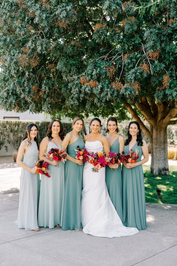 Bridesmaids standing in a line under a tree with bride all holding bouquets and smiling.