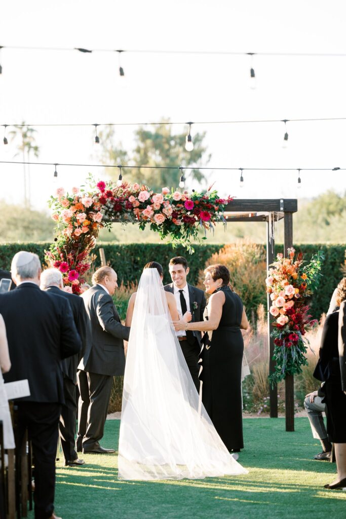 Bride being given away at wedding ceremony by mother and father with groom under chuppah.