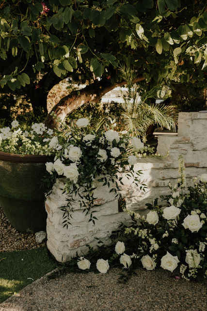 Wedding ceremony altar space flowers of white and greenery places on low wall and on ground.