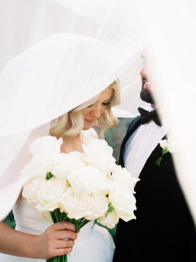 Bride and groom smiling under wedding veil flowing around them.