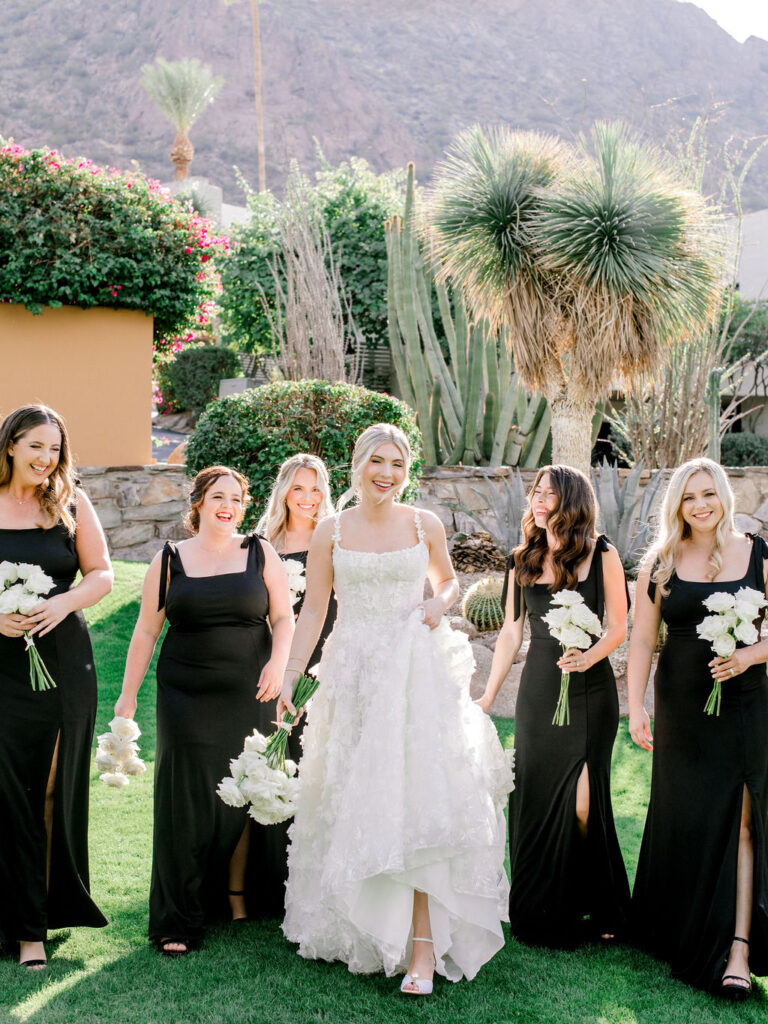 Bride walking with bridesmaids wearing black dresses, all holding white flowers bouquets, smiling.