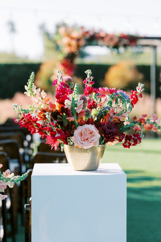 Back of wedding ceremony aisle floral arrangement in brass vase, sitting on white acrylic column.