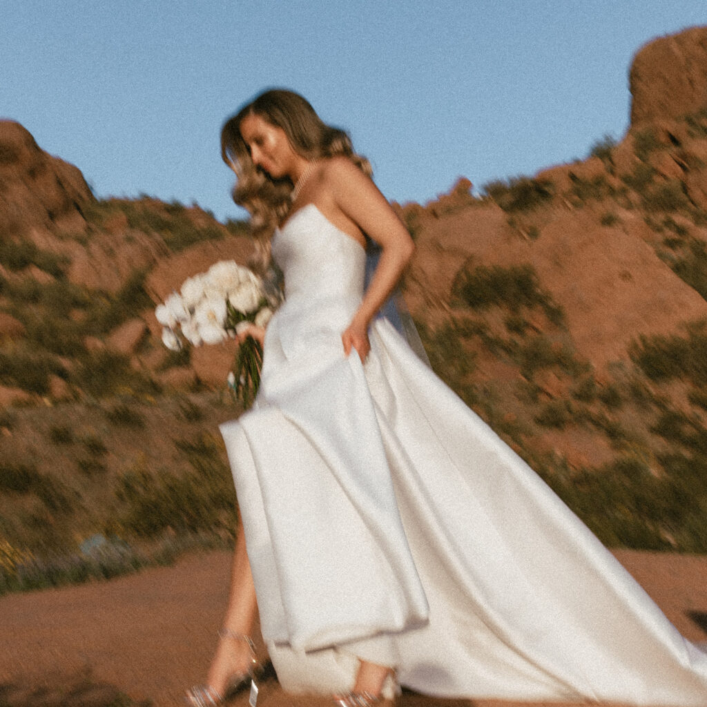 Blurry image of bride walking through dessert holding long stem roses bouquet.