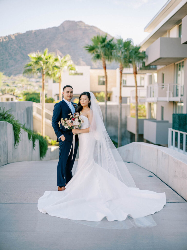Bride and groom standing next to each other and paved concrete walkway with no expressions on their face and desert landscape and building behind them.