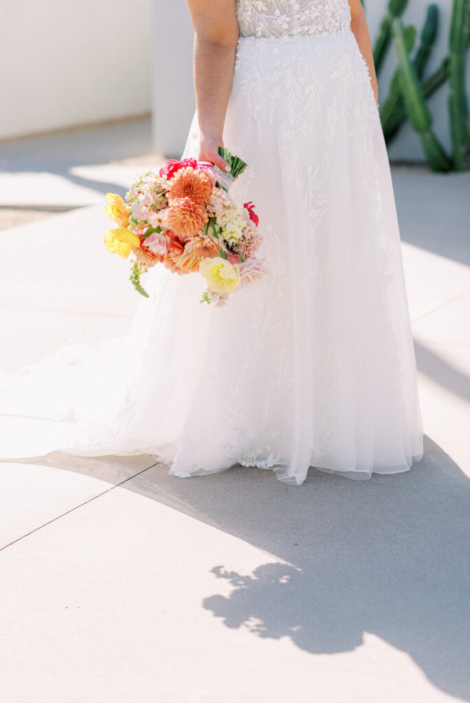 Bride holding bouquet of bright yellow, orange, pink, and cream colors, down at her side.