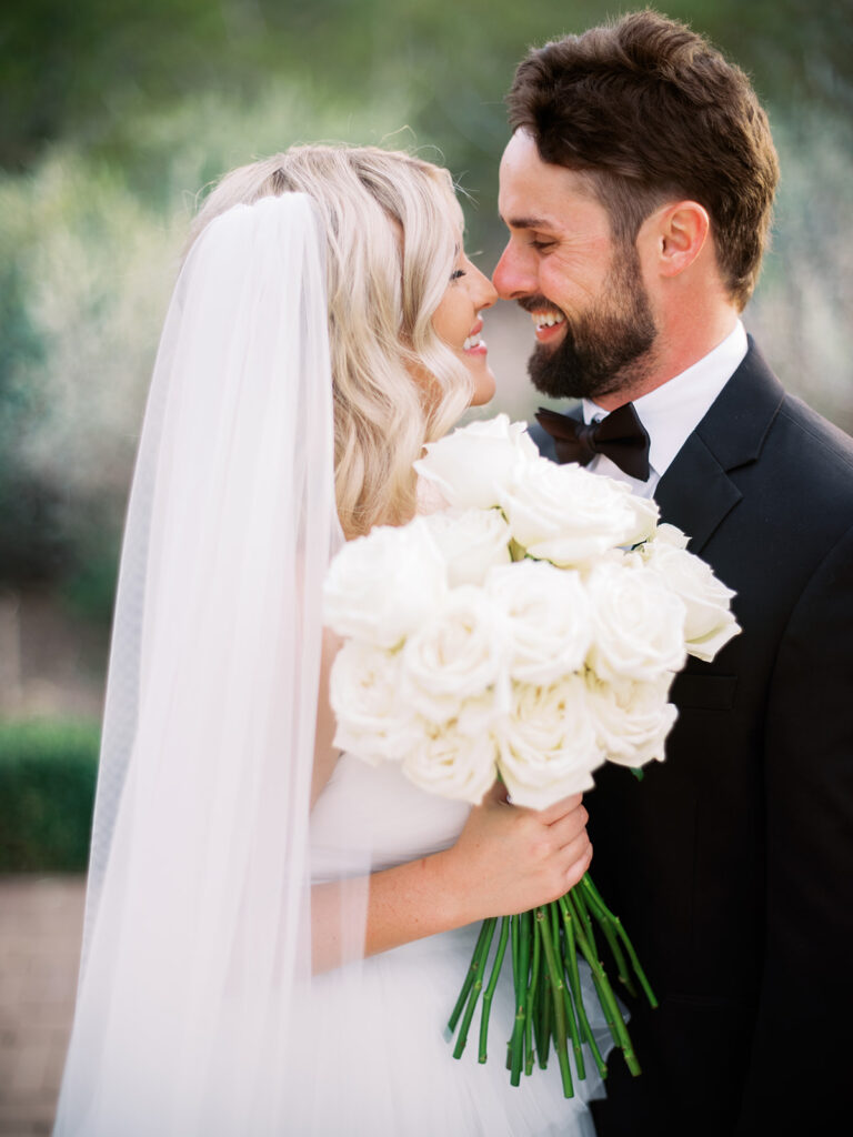 Bride and groom embracing, smiling, bride holding bouquet of white long stem roses.