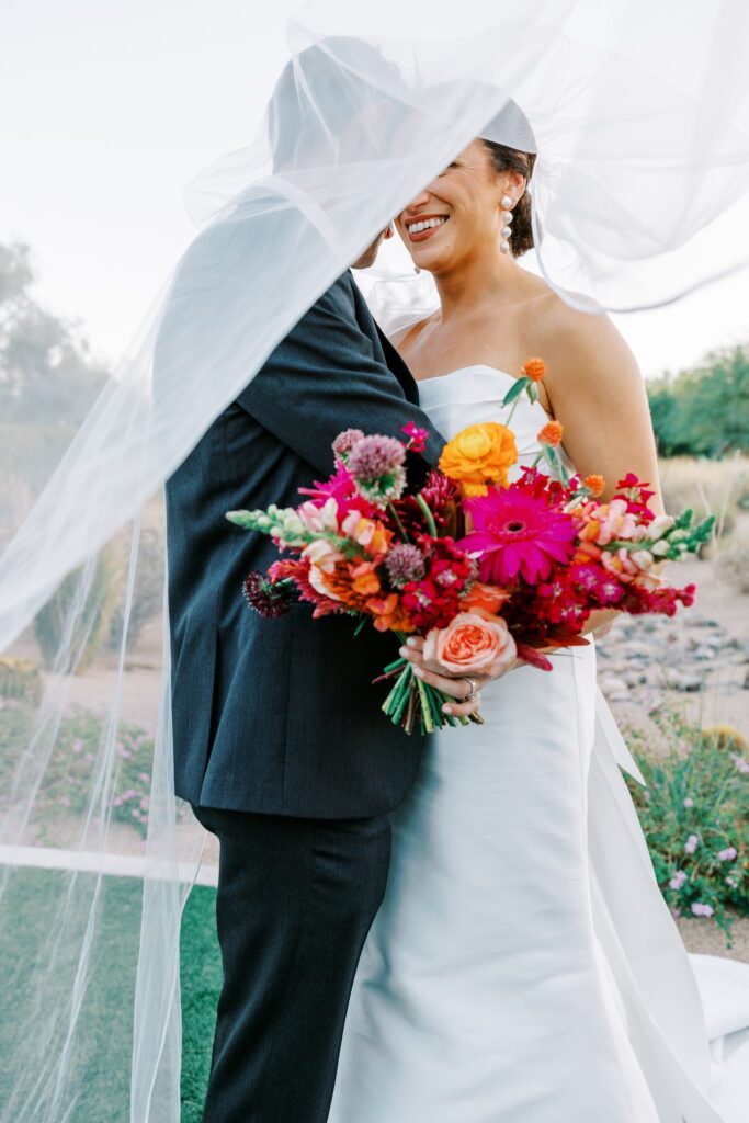 Bride and groom embracing under veil thrown over them, smiling, bride holding bouquet.