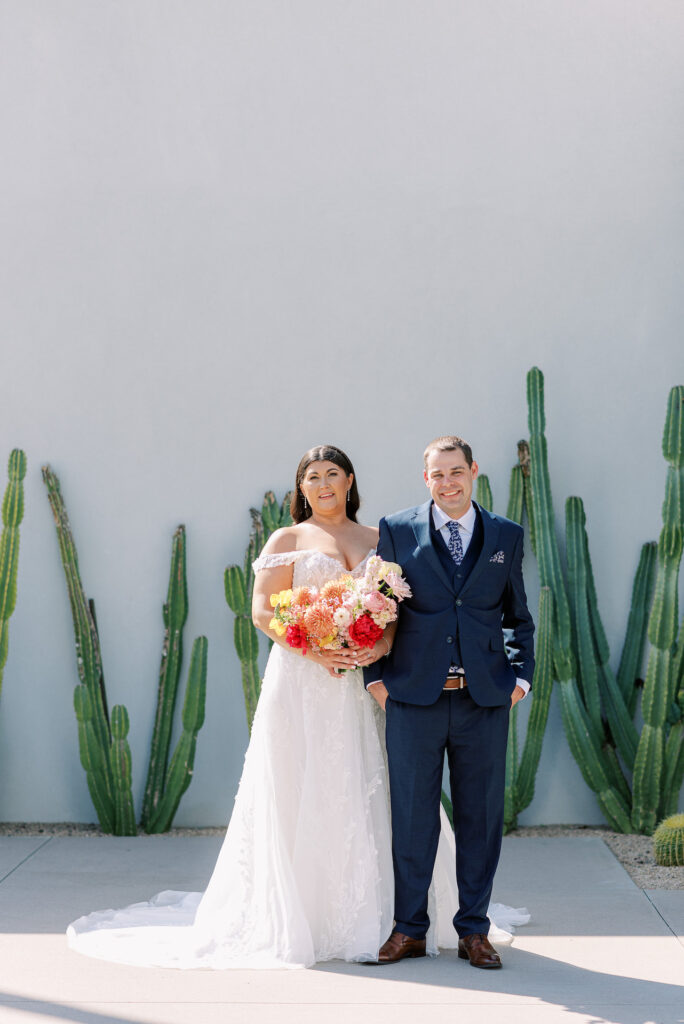 Bride and groom smiling, standing next to each other, bride holding bouquet, in front of white exterior wall with cacti growing in front of it.