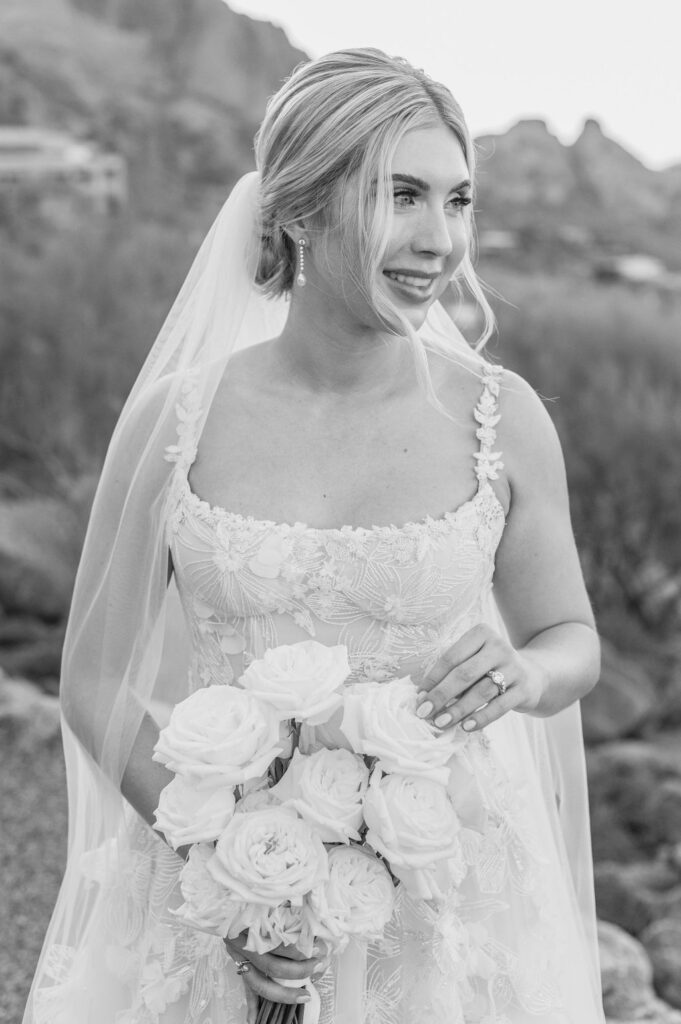 Bride holding bouquet of roses smiling and looking off to side in desert landscape.