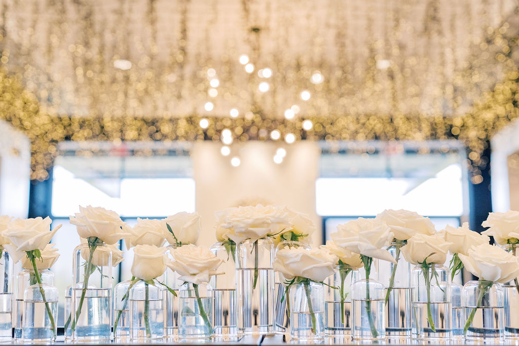 Many glass bud vases with white roses in them on a surface in room with gold sparkling on the ceiling.