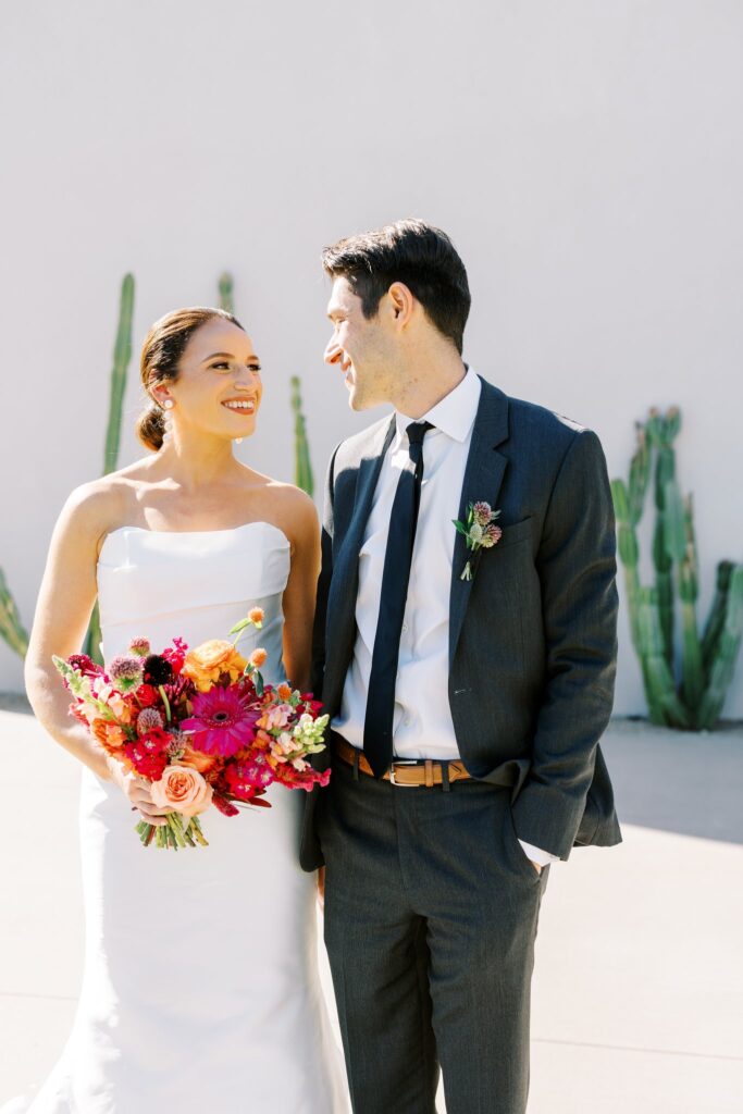 Bride and groom standing next to each other, looking at each other and smiling, bride holding bouquet.