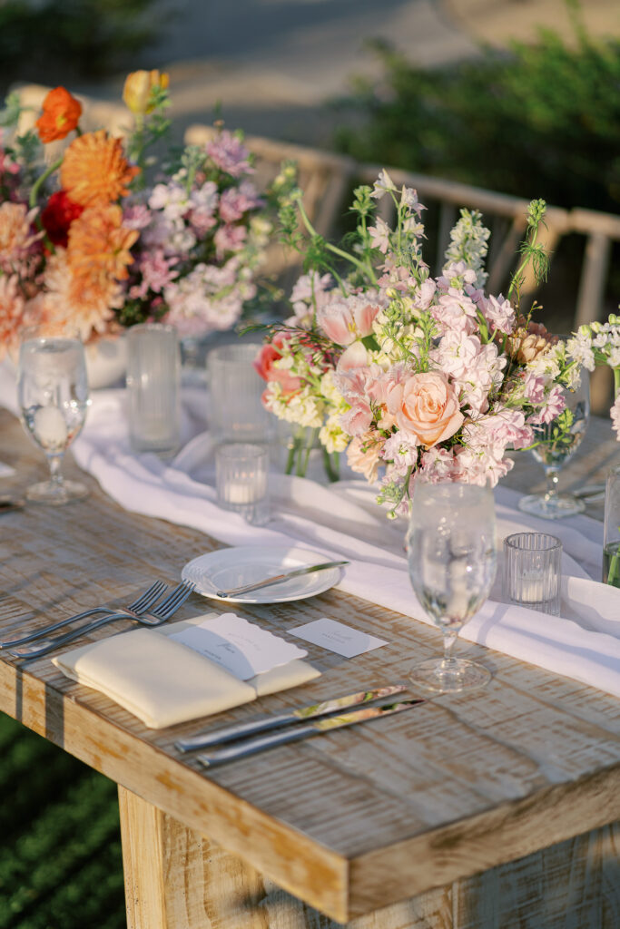 Long wood reception table with centerpieces down the middle placed on cloth with votive candles and flower centerpieces.