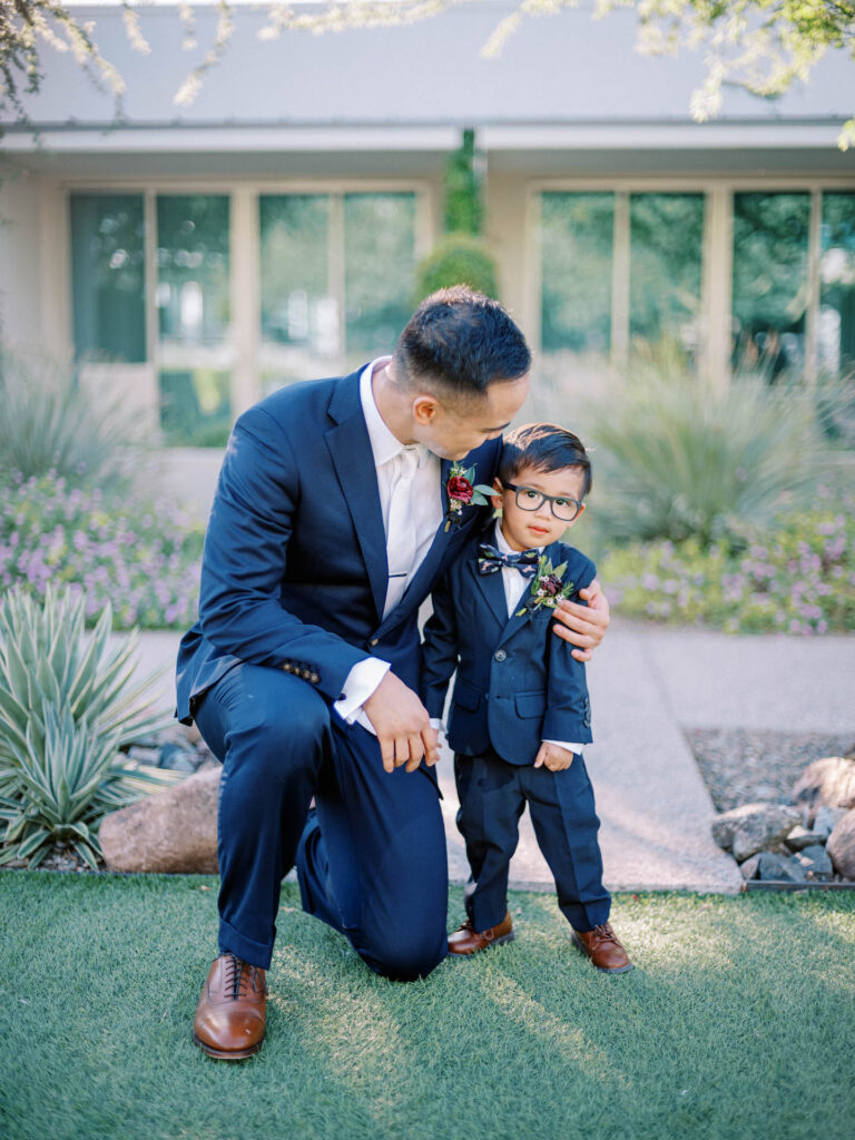 Groom kneeling down with his arm around and smiling at ring-bearer in blue suit.