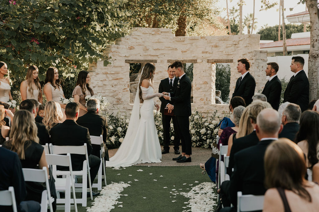 Bride and groom in altar space at outdoor wedding ceremony with guests seated and bridesmaids and groomsmen standing on either side.