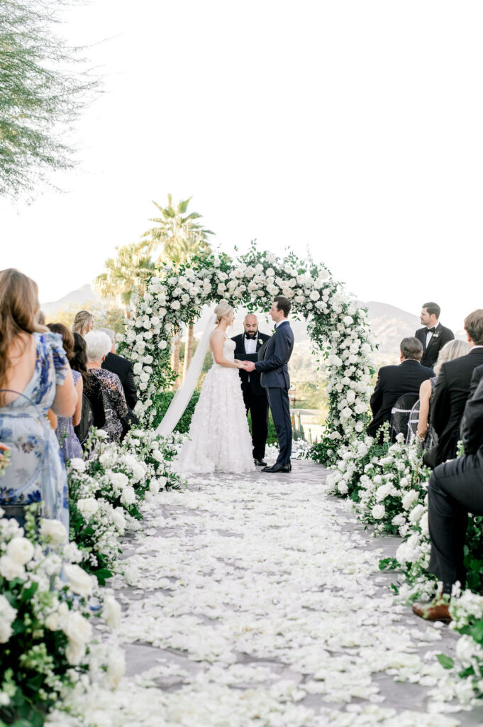 Bride and groom during outdoor wedding ceremony with officiant in altar space with a flower arch, aisle petals and aisle ground flower arrangements.