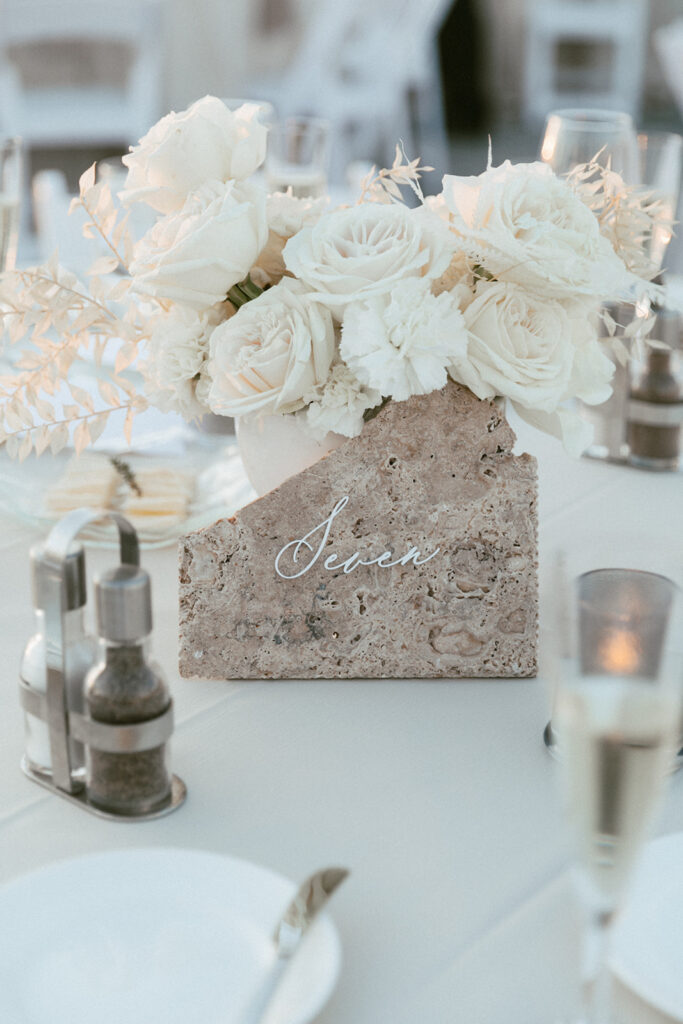 White roses and bleached Italian ruscus wedding reception centerpiece with stone table number sign in front.