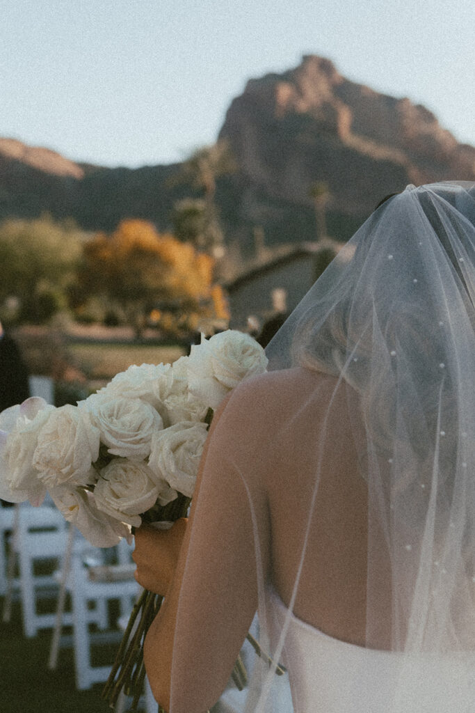 Image of bride standing while holding bouquet of long stem white roses with her veil covering he back.