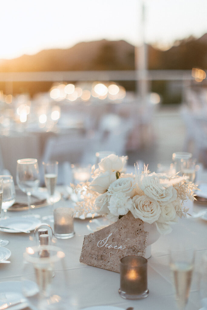 White roses and bleached Italian ruscus wedding reception centerpiece with stone table number sign in front.