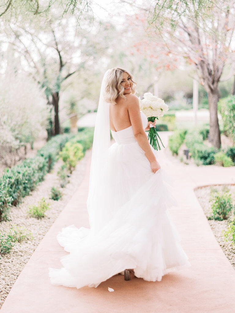 Bride walking down paved path smiling, looking back over her shoulder.