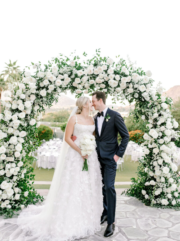 Bride and groom with their faces close under wedding ceremony arch, smiling.