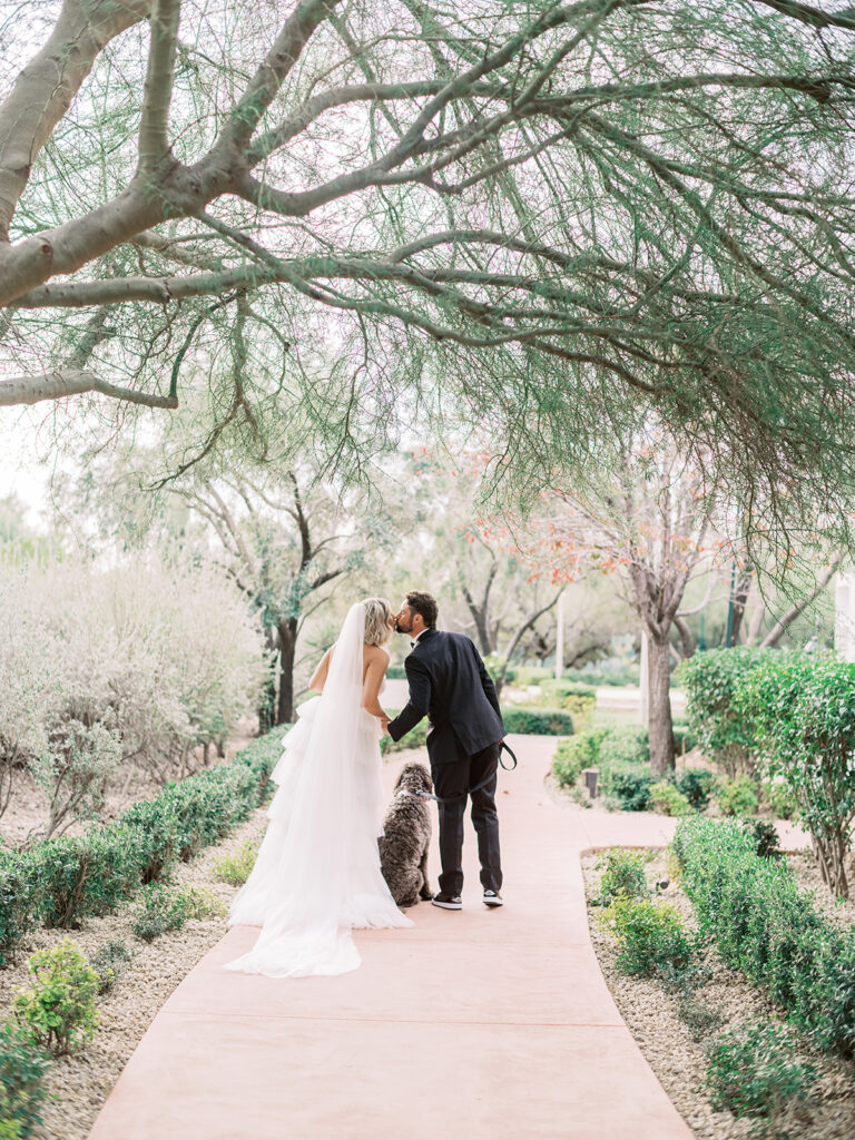 Bride and groom kissing on paved pathway with dog seated between them, all facing away.
