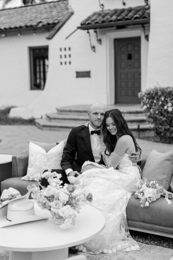 Groom smiling, bride embracing him and looking down, both seated on outdoor couch in front of Spanish style architecture building.