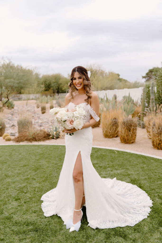 Bride standing in grass with desert landscape behind her, holding bouquet of white and blush roses.