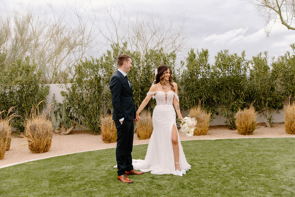 Bride and groom holding hands while standing in grass with desert landscape behind them.
