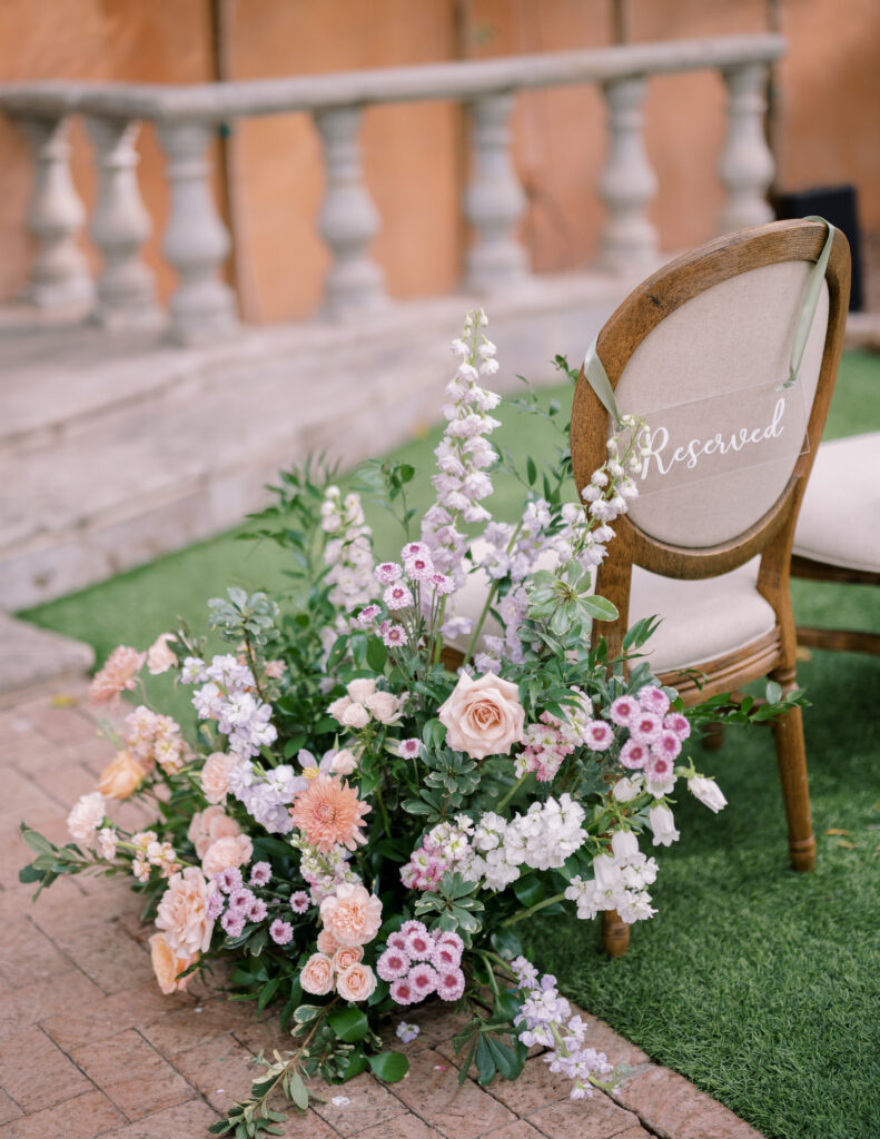 Ground floral arrangement next to aisle chair with purple, pink, and white flowers with greenery.