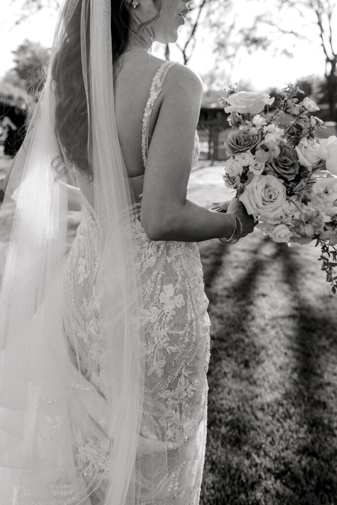 Black and white photo of bride's back holding bouquet and veil trailing down behind her.