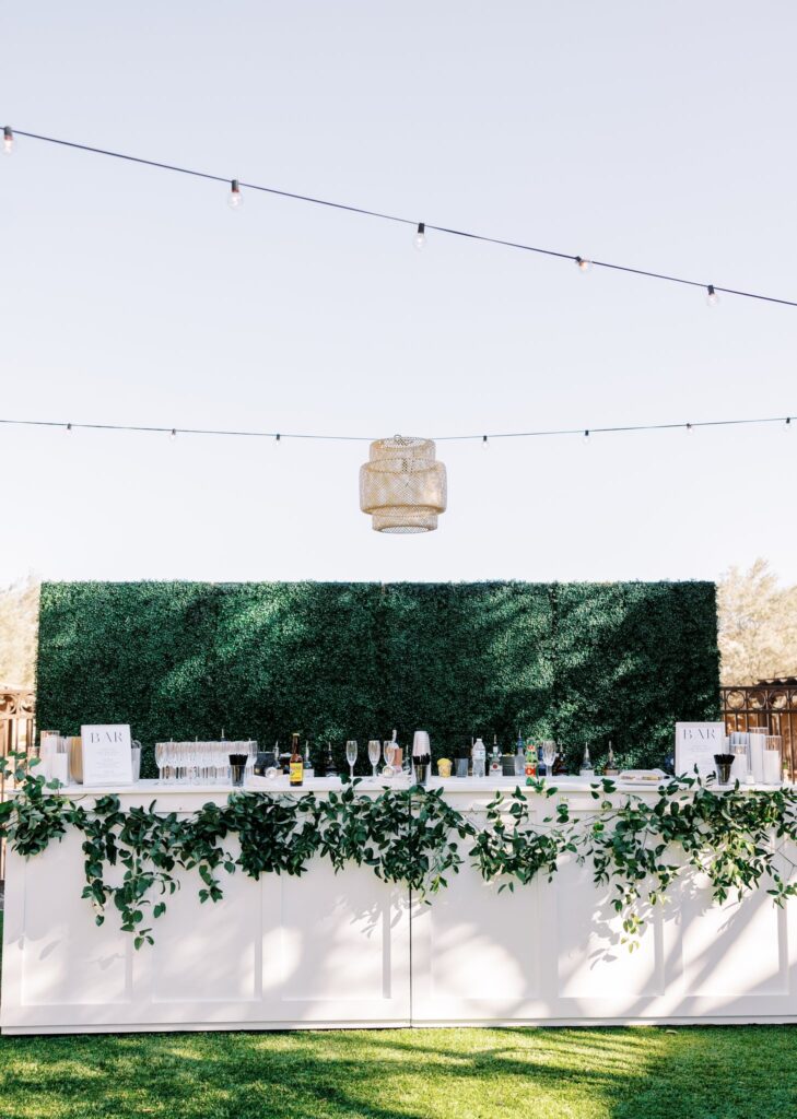 Greenery hanging from long white bar at wedding reception with greenery hedge behind it.