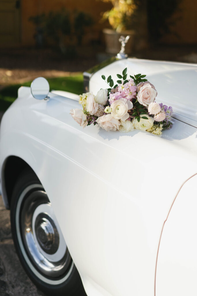 Bridal bouquet of pink, white, light purple, and cream flowers on hood of white fancy car.